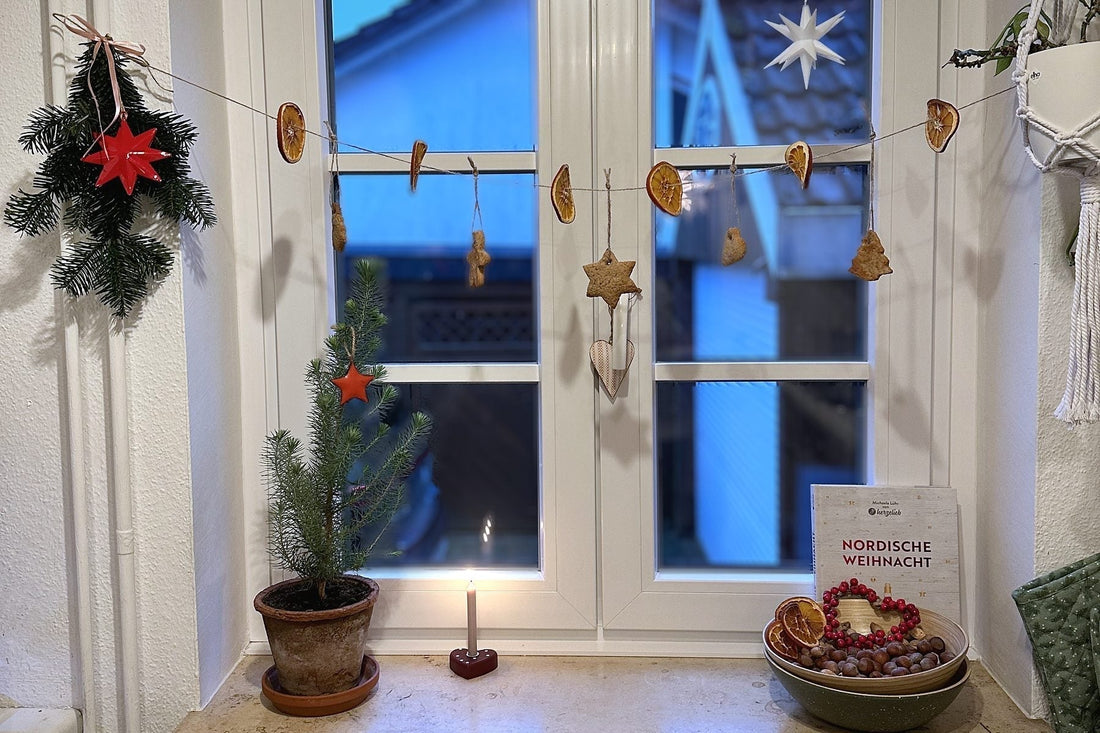 Küchenfenster in der Dämmerung mit Girlande aus Orangenscheiben und Plätzchen, kleiner Tanne im Tontopf und brennender Kerze