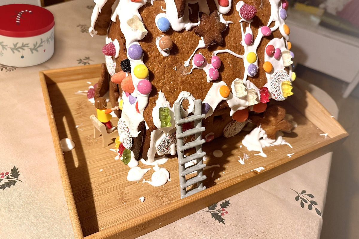 Selbst gebackenes Lebkuchenhaus mit Zuckerguss und bunten Süßigkeiten auf einem Holztablett, daneben eine Plätzchendose mit rotem Deckel