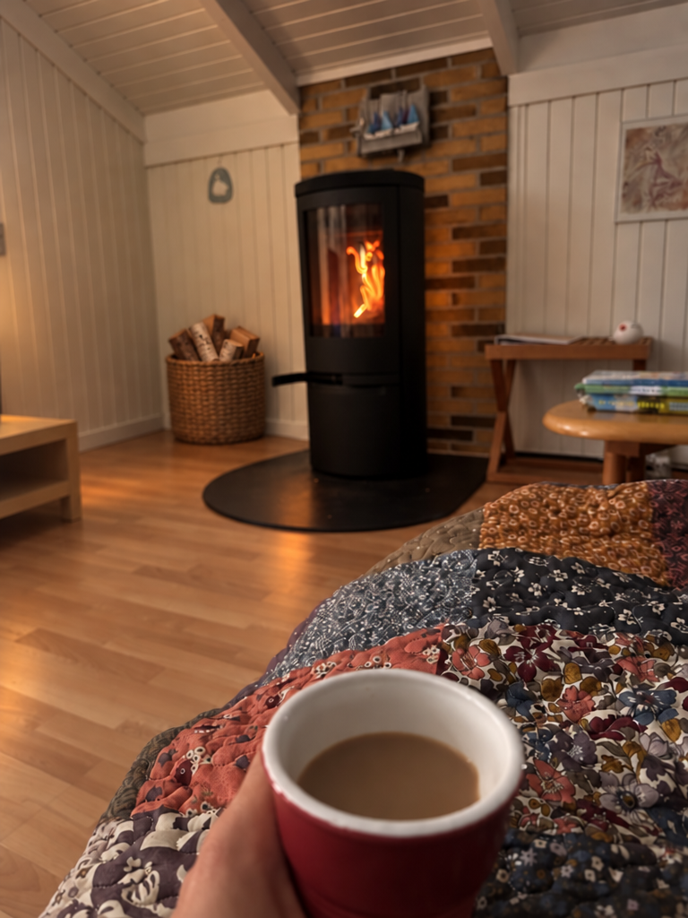 Kaminofen mit Feuer vor einer Ziegelwand in einem dänischen Ferienhaus, im Vordergrund eine Patchworkdecke und eine Tasse Kaffee