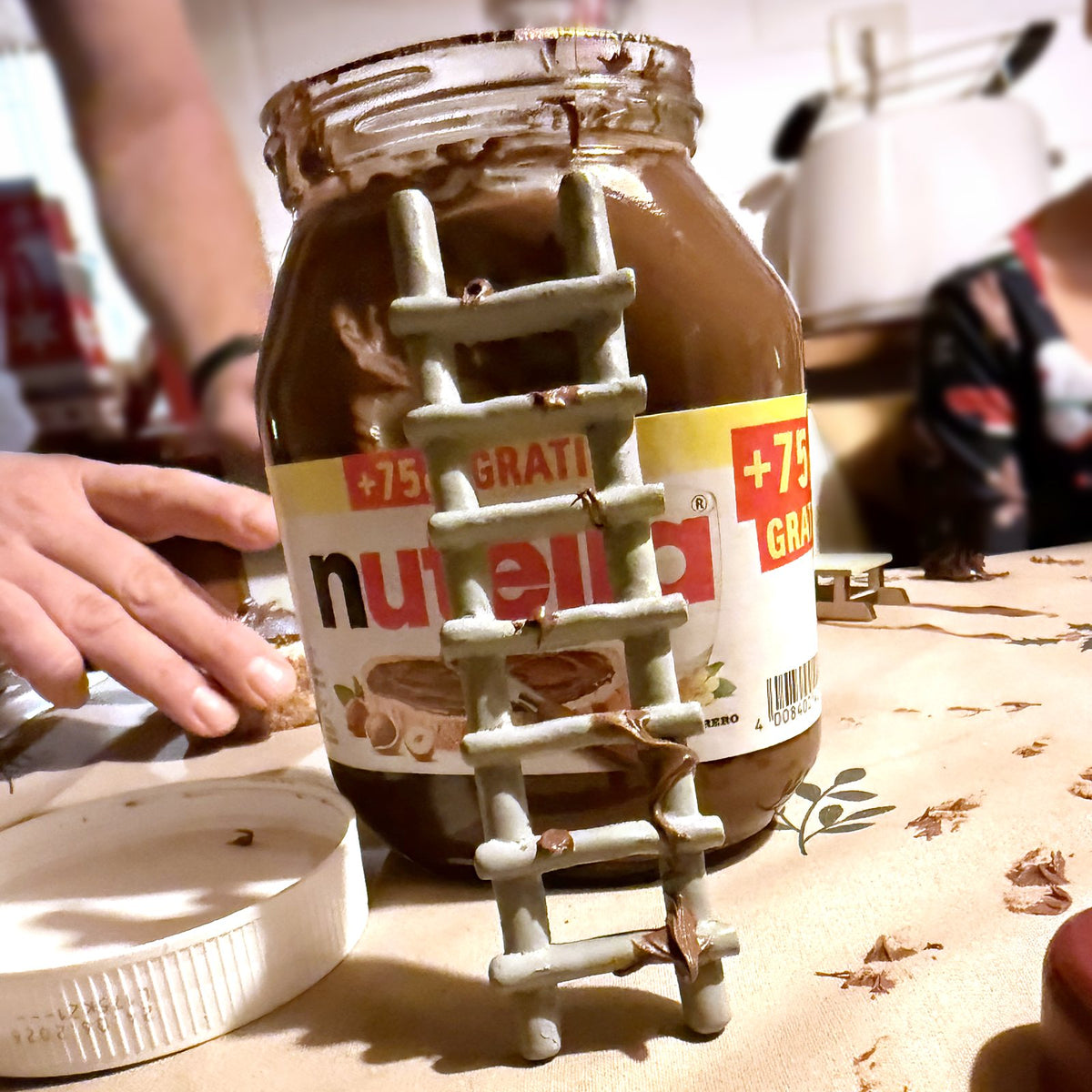 Kleine Wichtelleiter lehnt an einem offenen Glas Nuss-Nougat-Creme, auf dem Tisch Schokoladenspuren