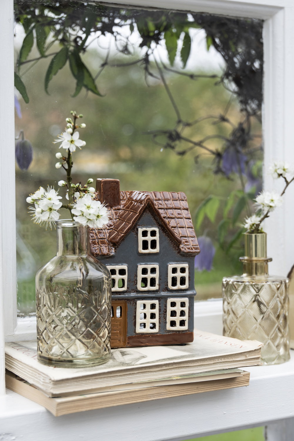 Graublaues Teelichthaus mit weißen Blüten in der Glasflasche auf der Fensterbank